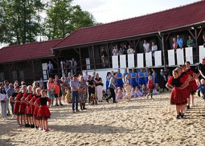 Zdjęcie przedstawia tancerki ustawione w pozach tanecznych. Tancerski stoją na piasku. W tle zabytkowa zabudowa Plaży Miejskiej oraz widzowie.