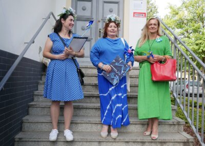 Zdjęcie przedstawia trzy kobiety stojące na schodach. Dwie z nich ubrane na niebiesko, mają na głowach wianki, trzymają w rękach podkładki do dokumentów. Trzecia ubrana na zielono, trzyma czerwoną torebkę. W tle szare drzwi.