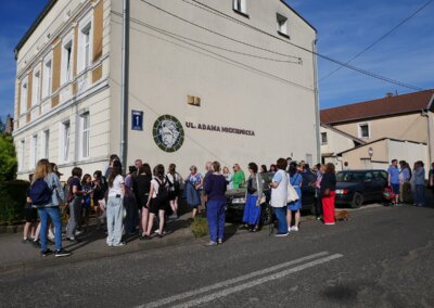 Zdjęcie przedstawiam grupę osób parzących na budynek, na którym zamieszczona jest ceramiczna postać Adama Mickiewicza wraz z napisem ul. Adama Mickiewicza. Powyżej napisu umieszczono ceramiczną otwartą książkę.