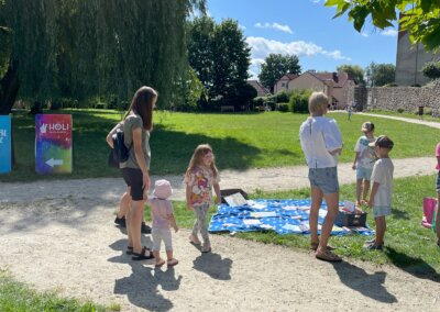 Dwie dorosłe kobiety i czwórka kilkuletnich dzieci stoją w parku wokół piknikowego koca, na którym leży kilkanaście książek.