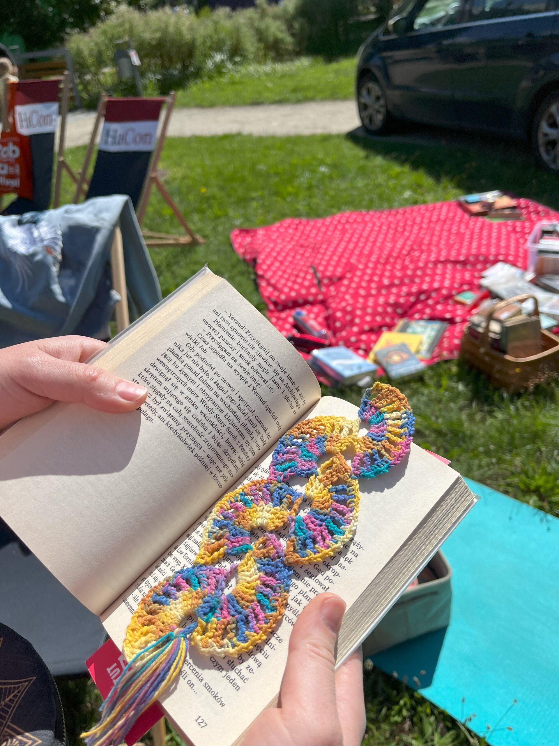 Ręce trzymają rozłożoną książkę, na której leży szydełkowa, pastelowa zakładka. W tle widać park z rozłożonym kocem piknikowym, leżaki i leżące książki.