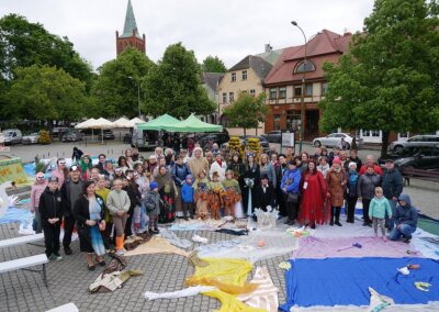 Grupa około 60 osób, w różnym wieku – dzieci, młodzież, dorośli i seniorzy – pozuje do wspólnego zdjęcia na barlineckim rynku. Uczestnicy stoją i siedzą wokół dużej, kolorowej instalacji przypominającej artystyczne jezioro, ułożonej z materiałów na brukowanej nawierzchni. Wiele osób ma na sobie barwne stroje inspirowane postaciami z legend i natury – widać m.in. skrzaty, Królową Puszczy Barlineckiej, boginkę Morianę, dzieci w maskach ptaków oraz kwiaty. W tle – zabytkowe kamienice, zielone drzewa oraz wieża kościoła z czerwonej cegły. Atmosfera jest radosna, mimo pochmurnego nieba i chłodnej pogody.