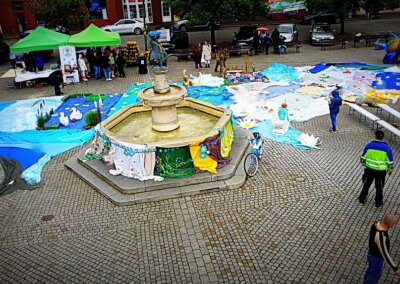 Zdjęcie wykonane z góry, ukazujące Rynek w Barlinku z kamienną fontanną w roli głównej. Fontanna ozdobiona jest kolorowymi materiałami przypominającymi kwiatową spódnicę – dzieło lokalnych artystek. Wokół niej rozpościera się rozległa, barwna instalacja z tkanin, imitująca jezioro, łąkę i postaci z legend – w tym łabędzie, lilie wodne oraz inne elementy przyrody. W tle widać zielone namioty z animacjami, ludzi zwiedzających oraz fragmenty zabudowy rynku. Po prawej stronie kadru rozstawione są ławki do siedzenia, a w tle – duży smok w kolorach tęczy.