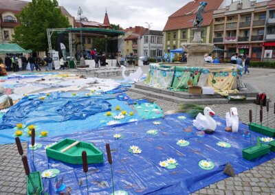 Zdjęcie przedstawia fragment barlineckiego rynku podczas plenerowego wydarzenia kulturalnego. Na pierwszym planie widoczna jest fontanna z rzeźbą Gęsiarki, udekorowana kolorowymi tkaninami przypominającymi kwietną spódnicę. Przed fontanną rozciąga się duża, barwna instalacja artystyczna z tworzyw i materiałów – niebieskie folie imitujące wodę, papierowe lilie wodne, trzcinowe elementy, makieta zielonej łódki, a także dwie rzeźby łabędzi. W tle znajduje się scena z nagłośnieniem, ludzie stoją przy stoiskach. Otoczenie to niskie budynki, sklepy i kamienice z czerwonymi dachami, a niebo jest pochmurne. Całość oddaje atmosferę rodzinnego, artystycznego święta inspirowanego naturą i lokalnymi legendami.