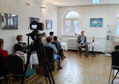 Na środku zdjęcia znajduje się prelegent Igor Kopa. Przy stolikach siedzą uczestnicy wydarzenia, za nimi na ścianie wiszą obrazy przedstawiające statki namalowane przez prelegenta. Widoczna jest również kamera na statywie, uwieczniająca wydarzenie.