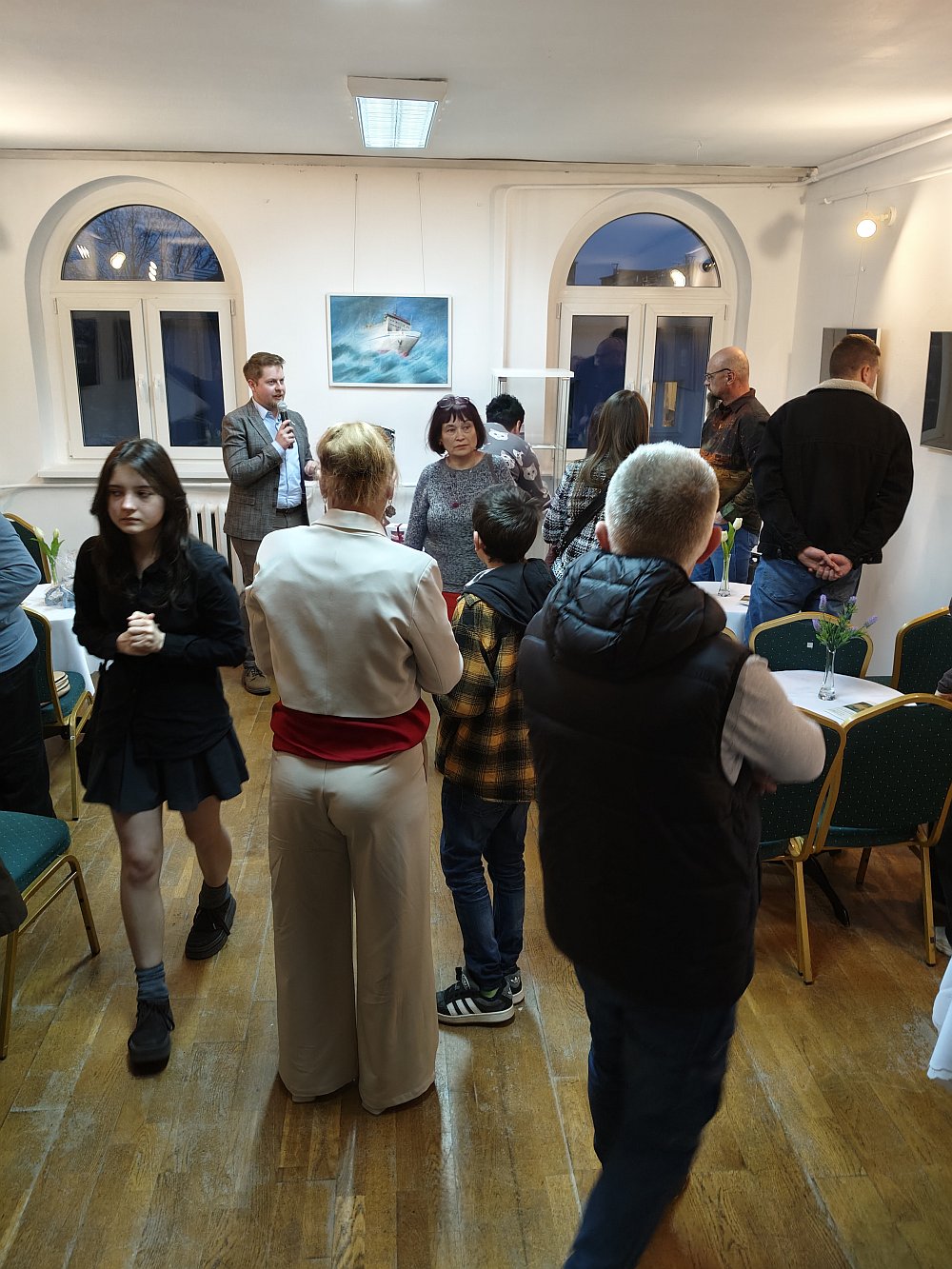Uczestnicy oglądający twórczość Igora Kopy. Na drugim planie prelegent opowiadający o statkach przedstawionych na obrazach. W tle gablotka z książkami o tematyce morskiej.