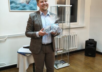 Prelegent Igor Kopa trzymający upominek, za nim znajduje się obraz przedstawiający prom „Jan Heweliusz” oraz gablotka z książkami i pocztówkami.