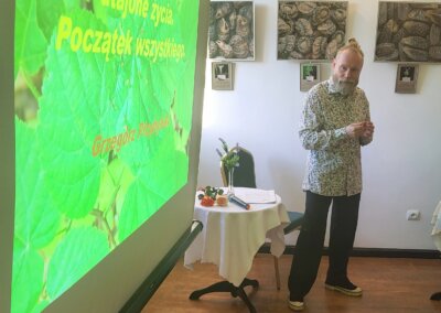 Prelegent Grzegorz Przybylski stoi obok ekranu z prezentacją zatytułowaną „Nasiona – utajone życie. Początek wszystkiego”. W tle na ścianach sali widoczne są prace z wystawy mikrofotografii nasion.