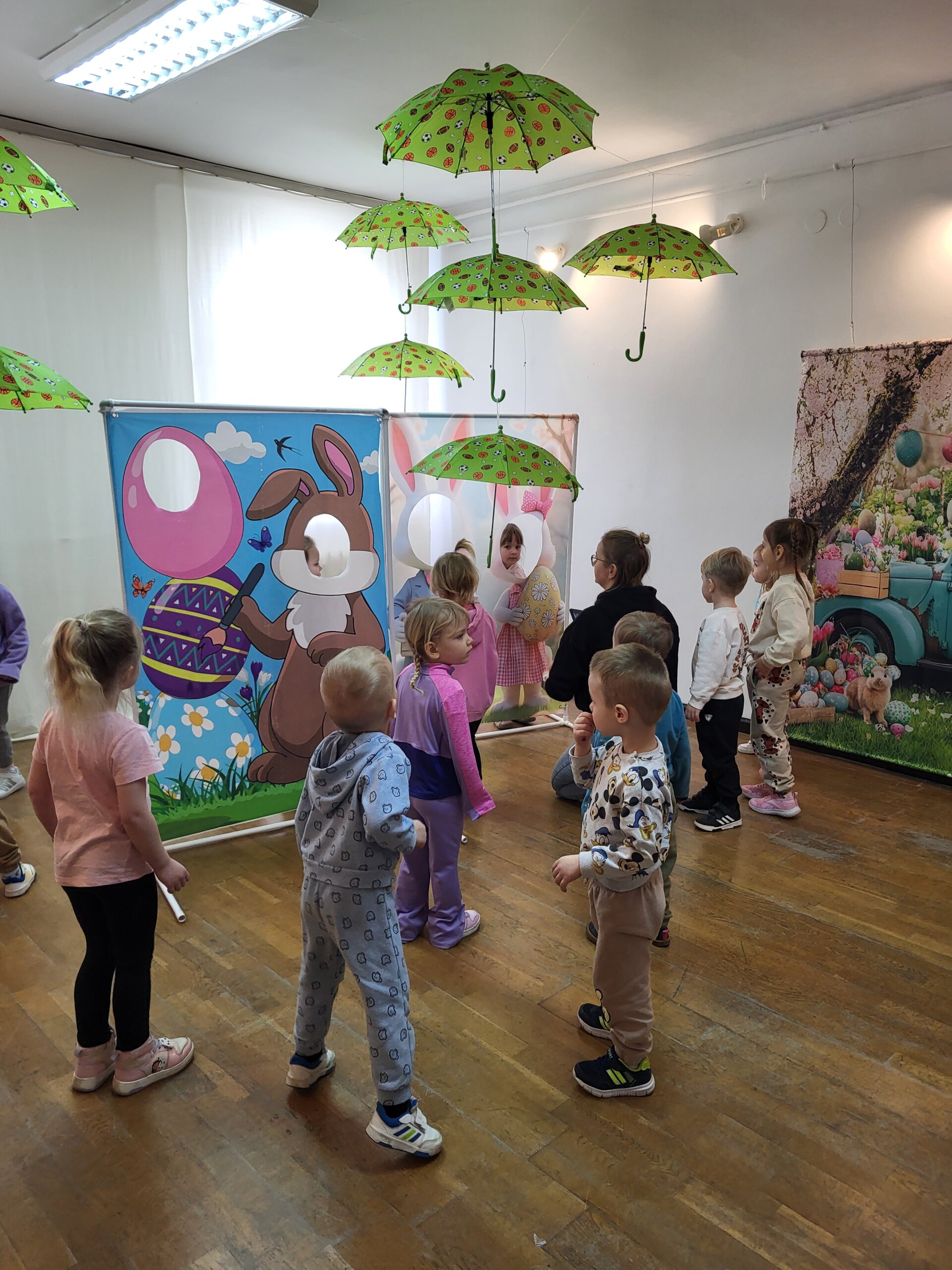 W Sali dzieci ustawione są tyłem do obserwatora, niektóre pozują przy wielkanocnych ściankach do zdjęć. Na ścianie widoczne jest kolorowe wielkanocne tło, a pod sufitem wiszą zielone parasole z wielkanocnymi wzorami.