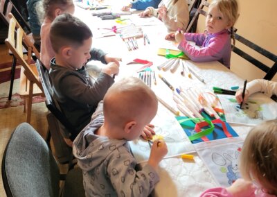 Grupa dzieci siedzi na krzesłach wokół podłużnego stołu i maluje wielkanocne torby z wizerunkami zajączków. Na stole leżą rozrzucone flamastry. W tle widać wiszący obraz na ścianie oraz zasłony w oknach.