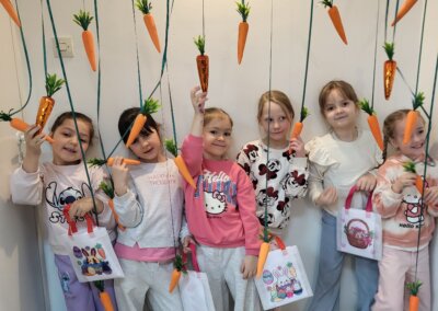 Grupa dziewczynek pozująca do zdjęcia na wielkanocnej ściance. Nad nimi wiszą dekoracyjne marchewki zawieszone na tasiemkach. Każda z nich trzyma w ręku marchewkę i uśmiecha się do zdjęcia.