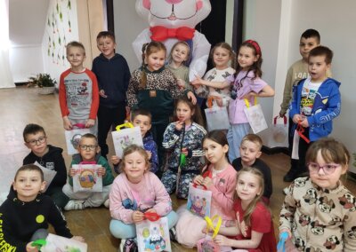 Zajączek wielkanocny stoi z grupą dzieci, które są ustawione wokół niego. Część z nich stoi inni siedzą na podłodze. W rękach trzymają kolorowe wielkanocne torebki. W tle na ścianie, wiszą dekoracje z kwiatów.