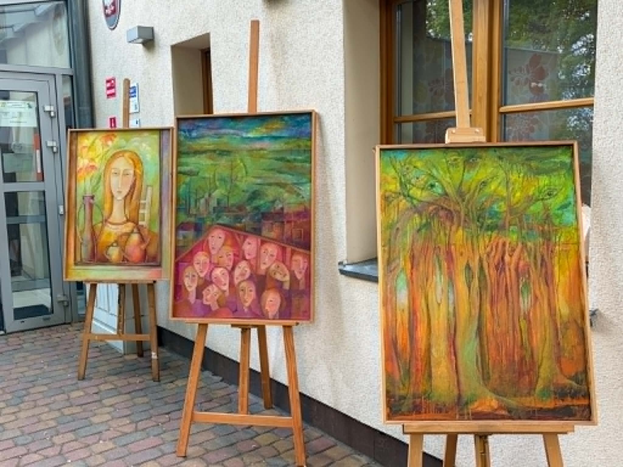 Wystawa na zewnątrz budynku, ustawione są trzy obrazy na drewnianych sztalugach. Obrazy są kolorowe i przedstawiają postać, pejzaż z grupą twarzy oraz las w ciepłych barwach. Przestrzeń jest otwarta, z szerokim przejściem umożliwiającym swobodne poruszanie się, także osobom korzystającym z wózków. Wejście do budynku znajduje się obok wystawy.