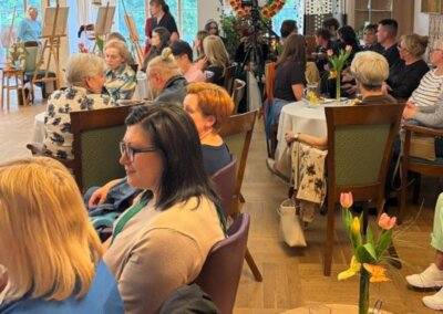 Spotkanie w sali, przestronnej sali odbywa się spotkanie artystyczne. Uczestnicy siedzą przy okrągłych stołach ustawionych z zachowaniem przejść między nimi. Na stołach znajdują się kwiaty i napoje. W tle widoczne są sztalugi z obrazami oraz duże okna zapewniające naturalne światło. Układ sali umożliwia łatwe poruszanie się i dostęp do miejsc siedzących.