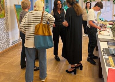 Wnętrze galerii / strefa rozmów. W pomieszczeniu galerii kilka osób stoi i rozmawia w pobliżu obrazów oraz stołu z materiałami informacyjnymi. Obrazy ustawione są na sztalugach na wysokości umożliwiającej oglądanie także osobom siedzącym. Pomieszczenie jest dobrze oświetlone i uporządkowane.
