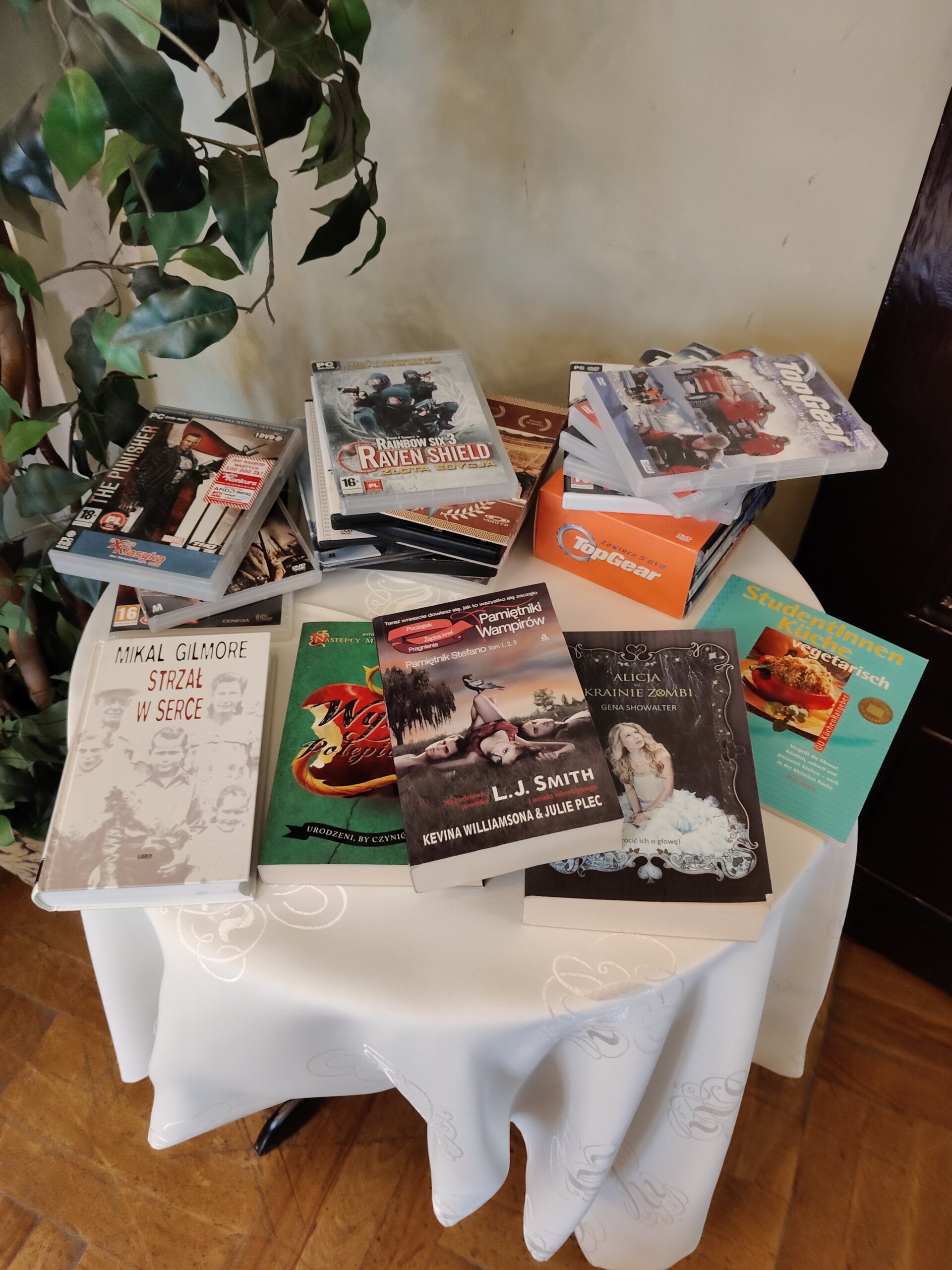 Na stoliku przykrytym białym obrusem leżą książki oraz filmy i gry na płytach przeznaczone do swobodnego zabrania przez uczestników wydarzenia.