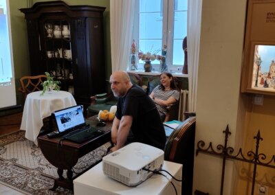 Prowadzący siedzi przy stoliku z laptopem podczas spotkania. W tle widoczna jest sala muzealna, ekran projekcyjny oraz uczestniczka wydarzenia.